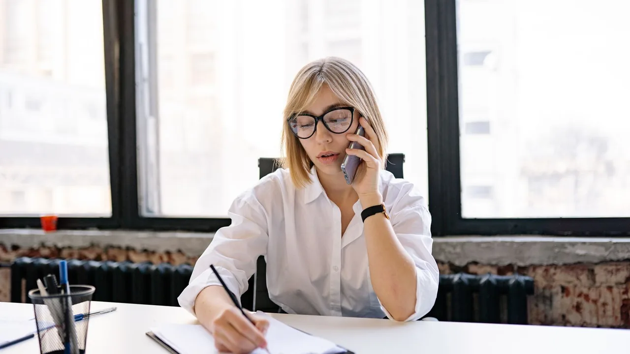 Une femme à lunettes téléphone à son bureau en prenant des notes, devant de grandes fenêtres