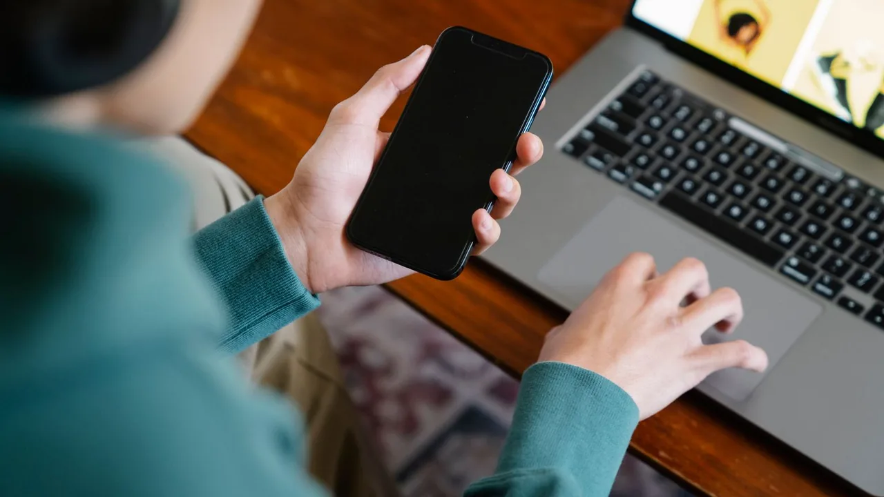 Une main tient un smartphone pendant que l'autre manipule le pavé tactile d'un ordinateur portable