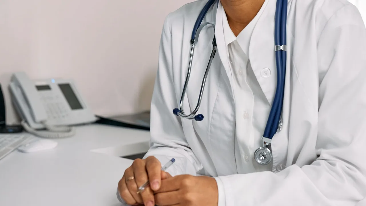 Médecin en blouse blanche et stéthoscope, mains jointes tenant un stylo, assis à un bureau avec un téléphone