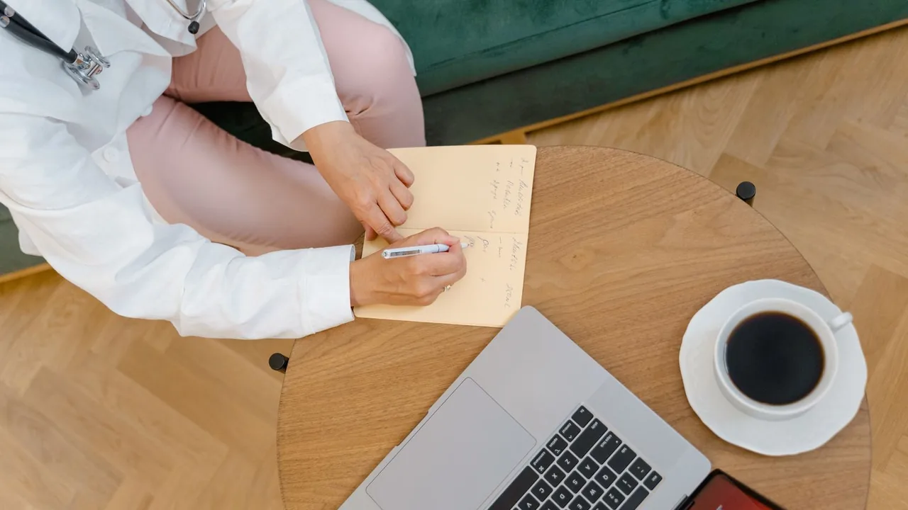 Vue de dessus d'un médecin prenant des notes dans un carnet, avec café et ordinateur portable sur une table ronde