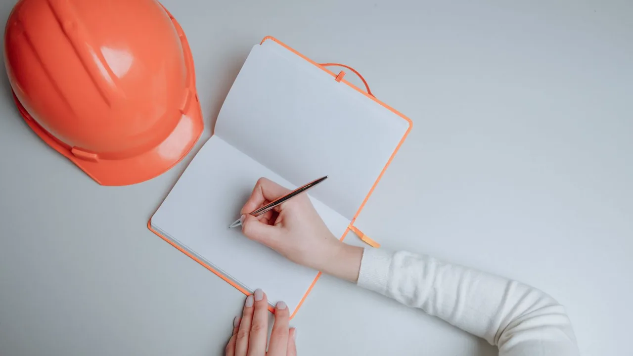 Vue de dessus d'une main qui écrit dans un carnet ouvert, à côté d'un casque de chantier orange