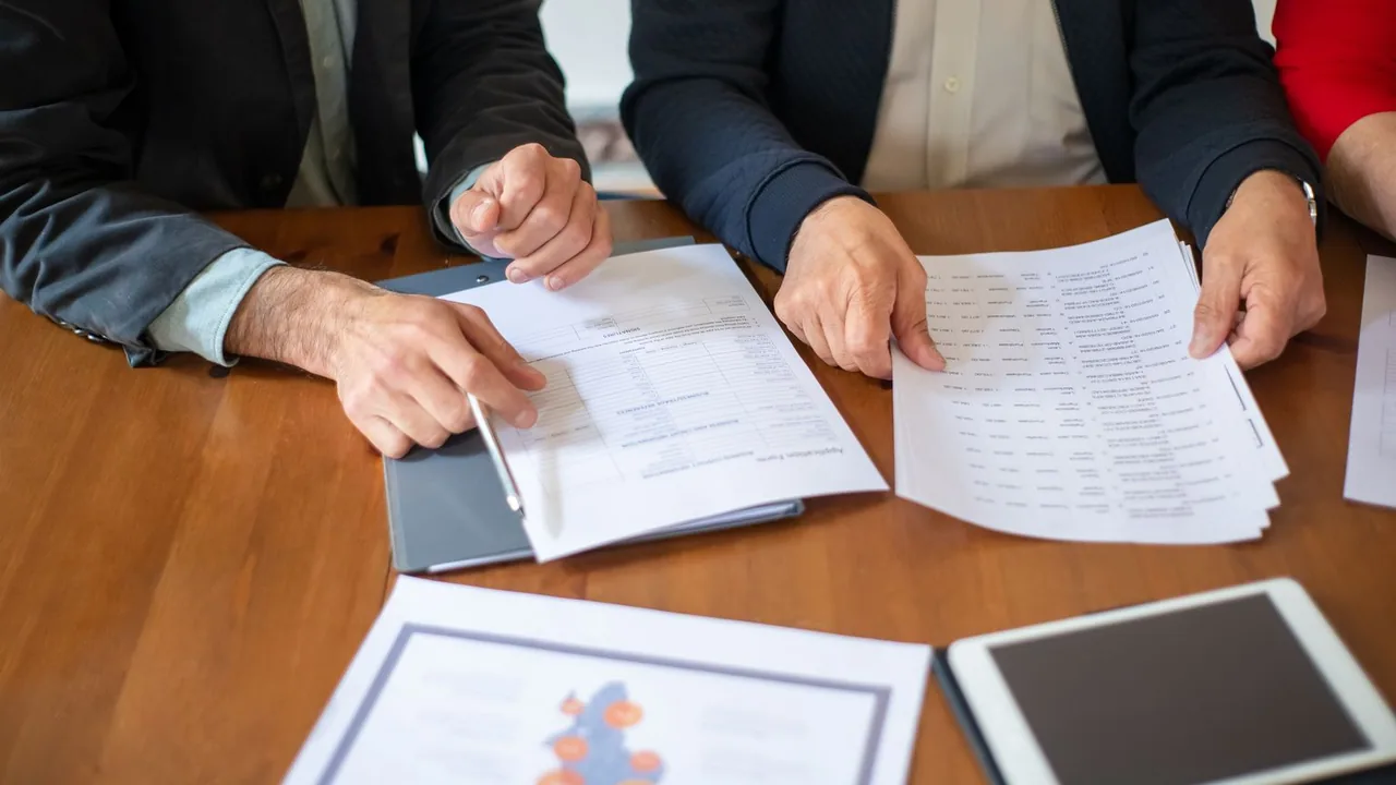 Deux professionnels comparent des tableaux de chiffres imprimés posés sur une table en bois, vue de dessus