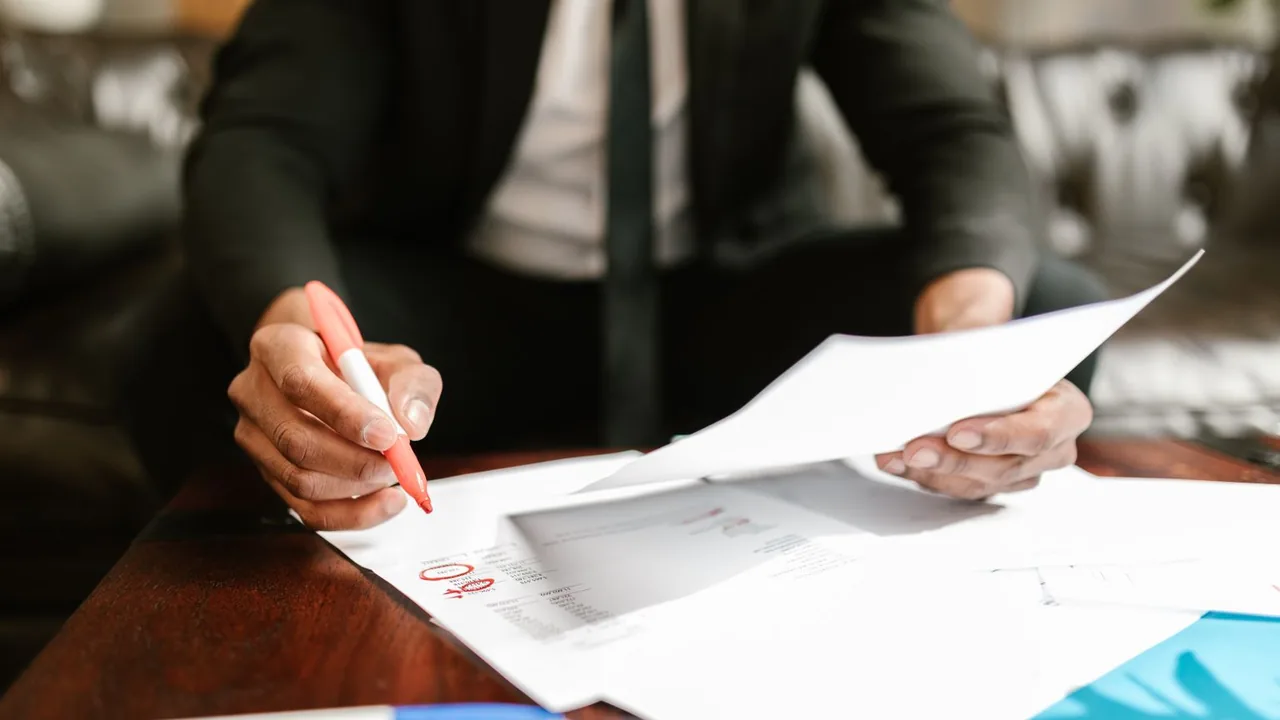Un homme en costume annote au stylo rouge des documents financiers imprimés posés sur un bureau