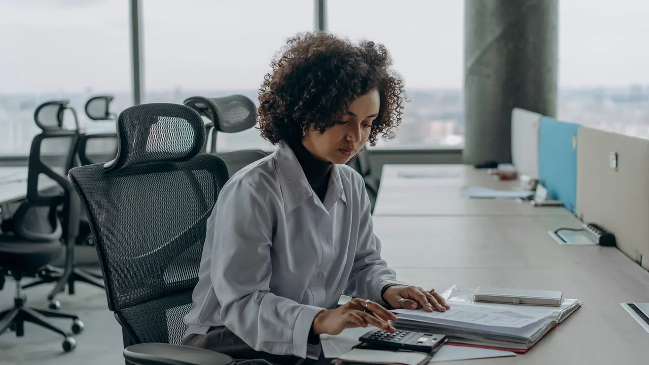 Une femme aux cheveux bouclés calcule des chiffres sur des documents à son bureau, devant de grandes fenêtres