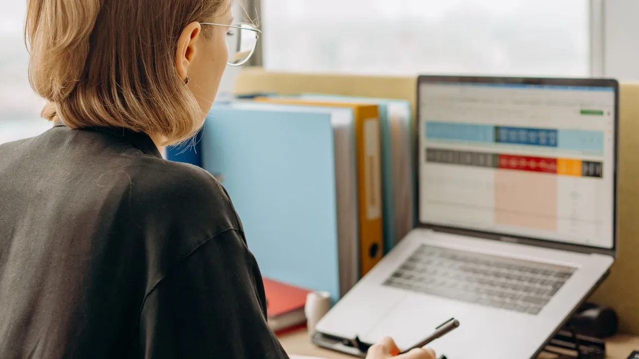 Une femme vue de dos consulte un tableau de données coloré sur son ordinateur portable, des classeurs à côté