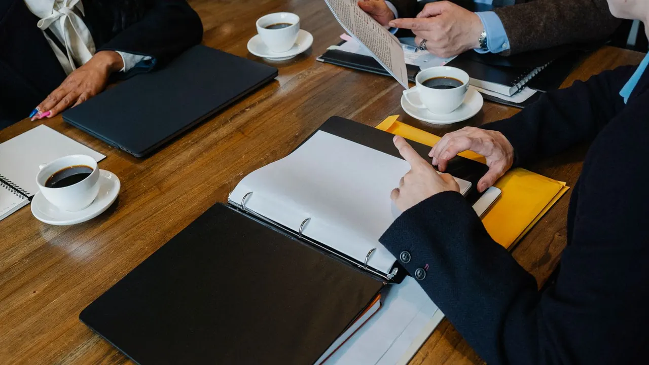 Des participants autour d'une table de réunion en bois, avec un classeur ouvert et des tasses de café