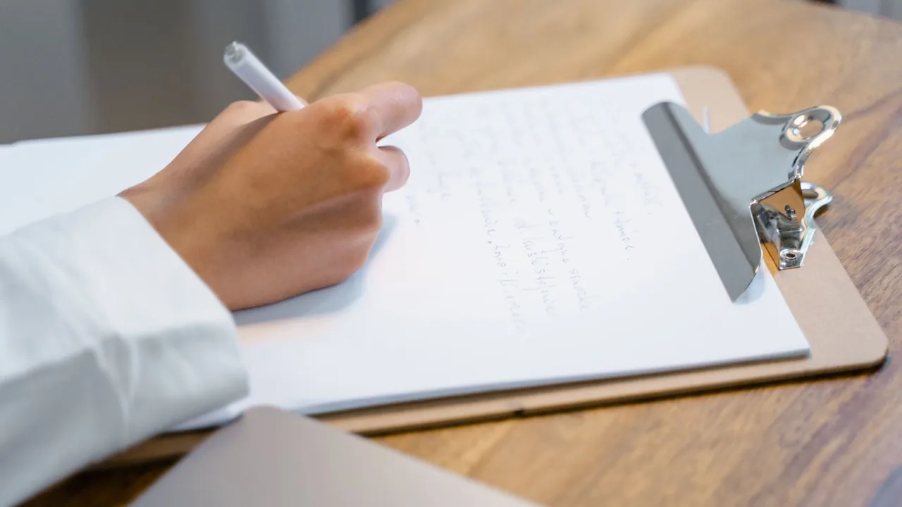 Une main écrit sur un bloc-notes à pince posé sur une table en bois