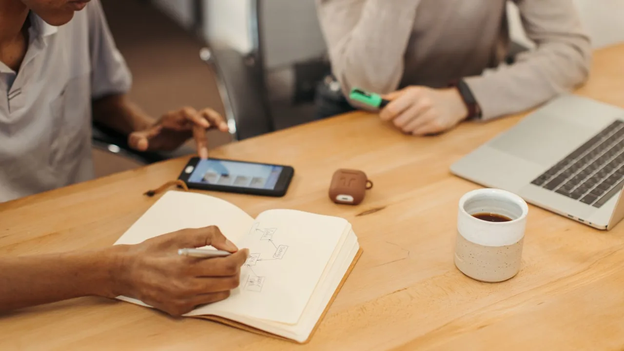 Une main dessine un schéma dans un carnet ouvert, avec un téléphone et un café posés sur la table