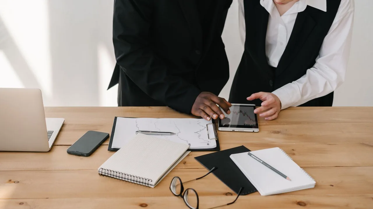 Deux personnes debout consultent une tablette affichant un graphique, carnets et ordinateur posés sur une table en bois