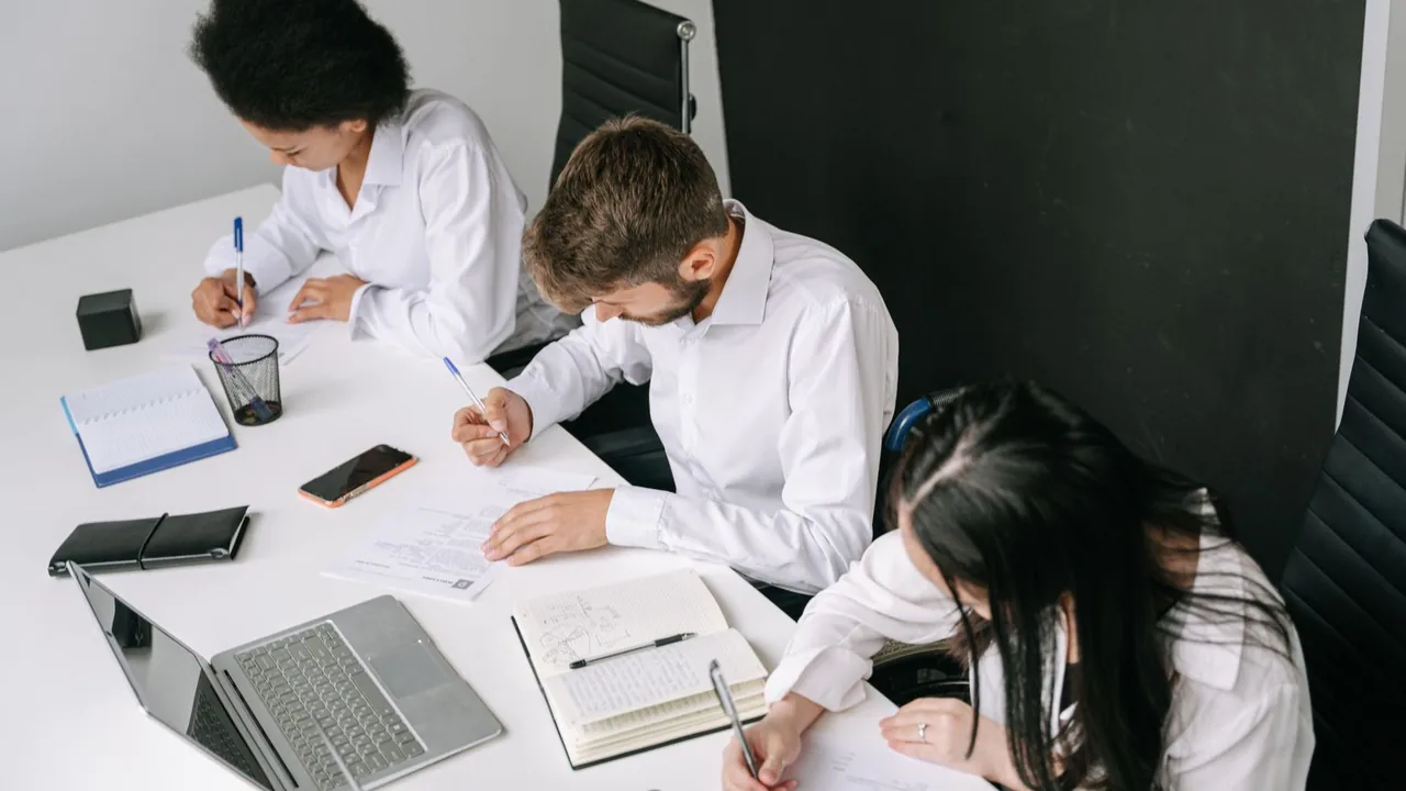 Trois collègues prennent des notes autour d'une table de réunion blanche, carnets et ordinateur ouverts