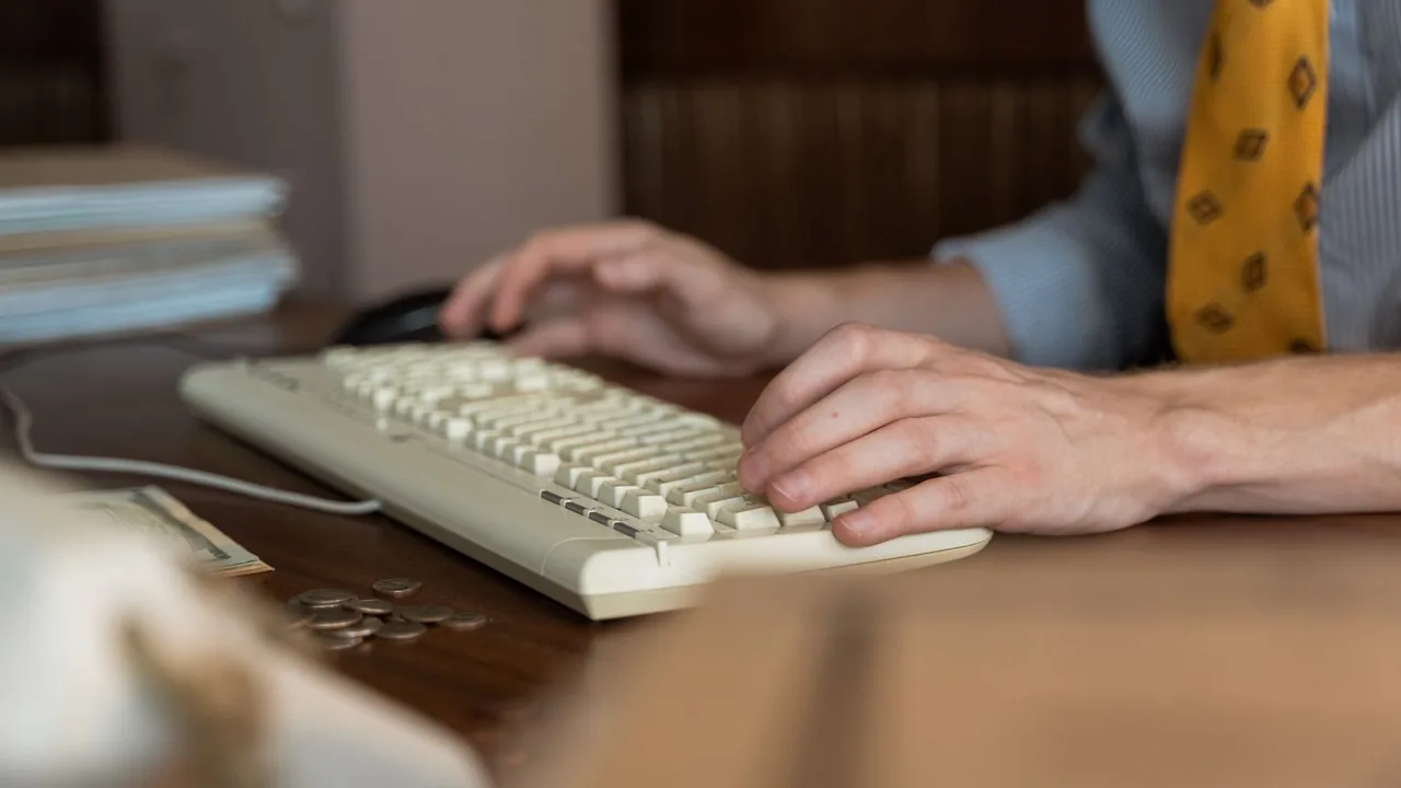 Mains d'un homme en cravate tapant sur un clavier, pièces de monnaie et dossiers sur le bureau