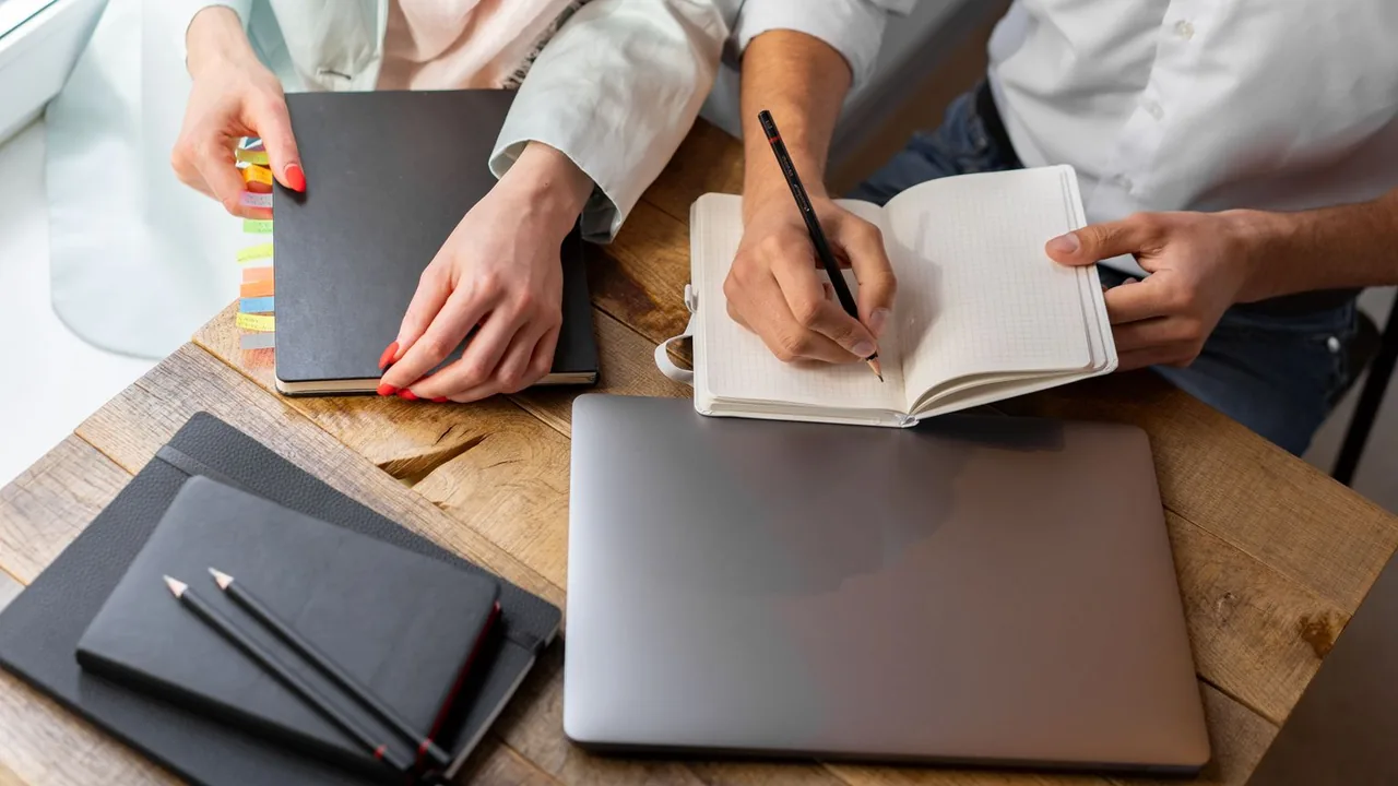 Mains écrivant dans un carnet à côté d'un ordinateur portable fermé et de carnets noirs sur une table en bois