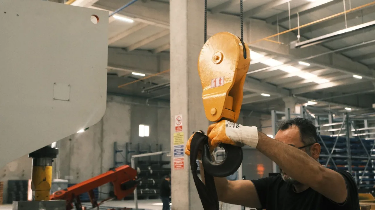 Ouvrier manipulant un crochet de levage jaune dans un grand hall industriel
