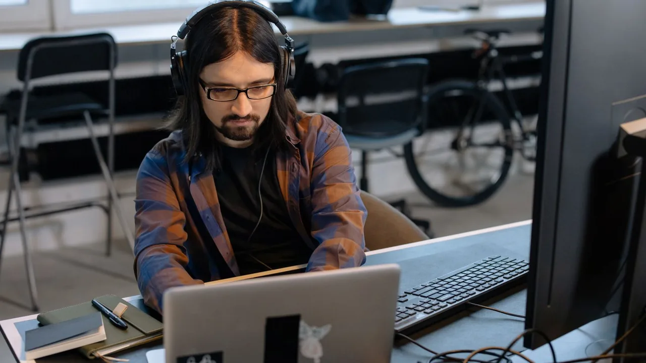 Un développeur barbu, casque sur les oreilles, code sur un ordinateur portable dans un open space