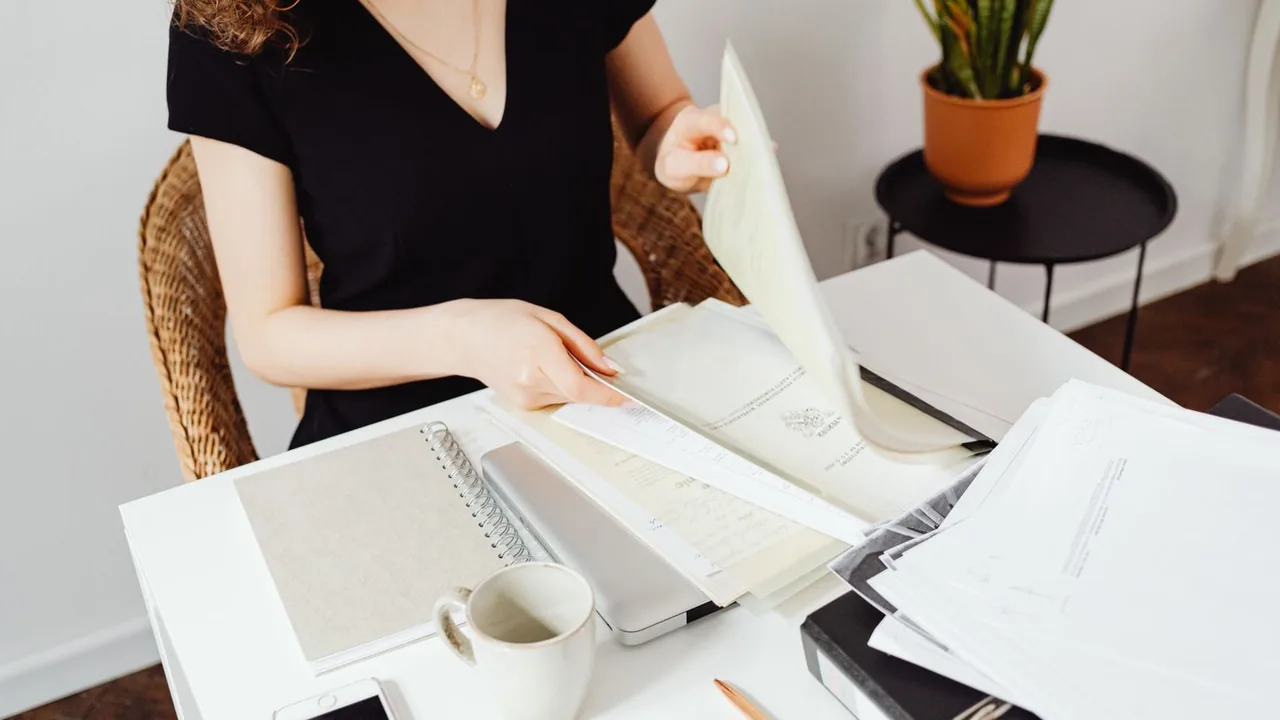 Une femme trie des documents imprimés à une table blanche, avec une plante et une tasse à côté d'elle.