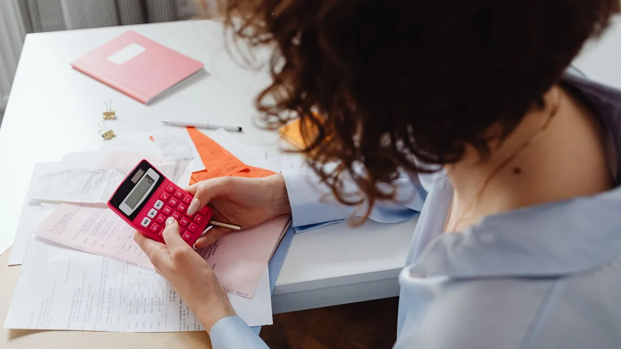 Une femme calcule des dépenses sur une calculatrice rose, entourée de factures et de reçus sur un bureau