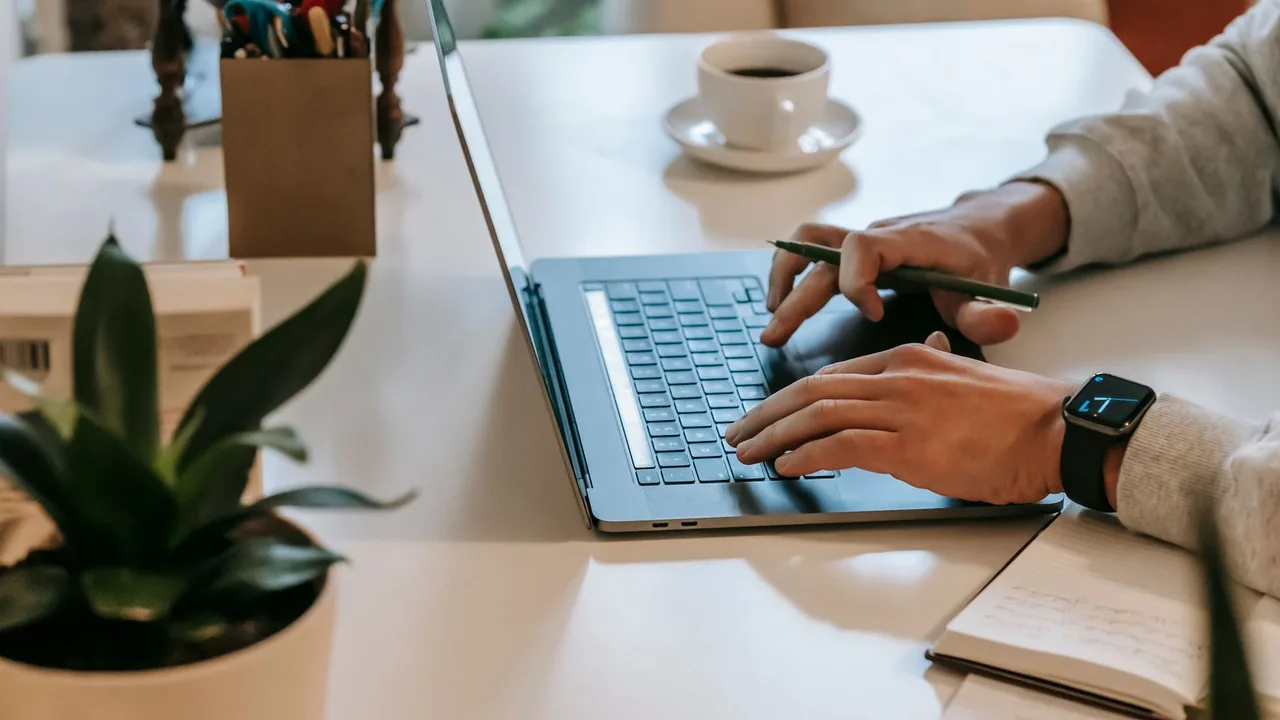 Mains tapant sur un ordinateur portable posé sur un bureau blanc, avec un café, une plante et un carnet
