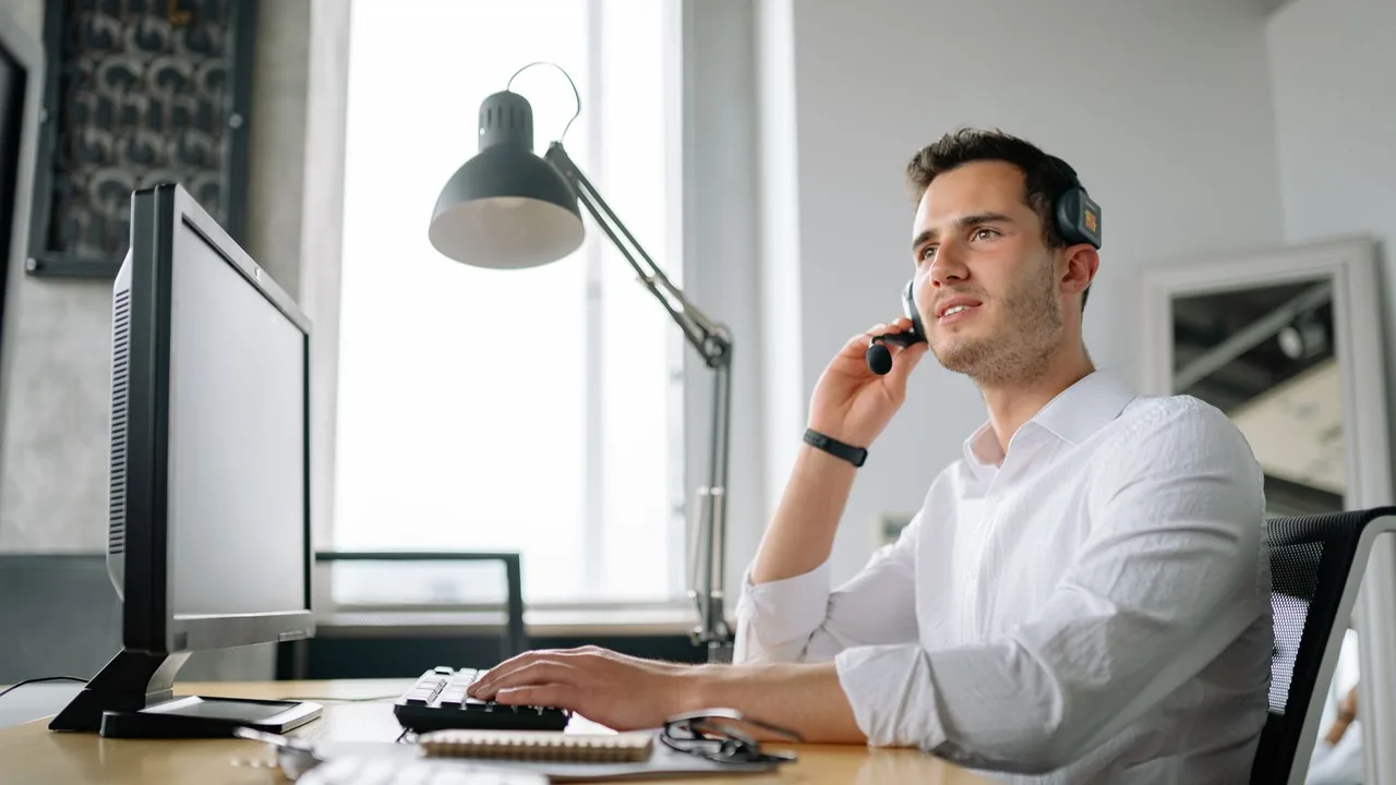 Un conseiller en chemise blanche, casque-micro sur l'oreille, travaille à son bureau devant un grand écran