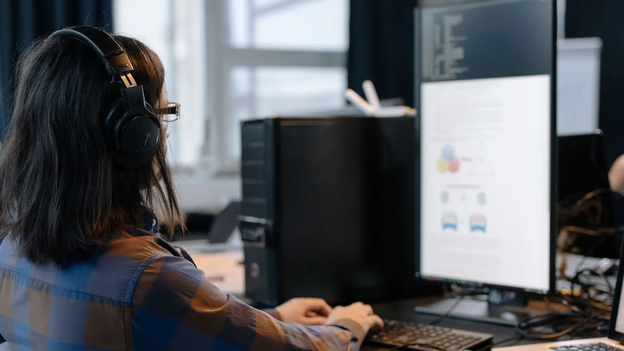 Une personne casque audio sur les oreilles, vue de dos, travaille au clavier devant un grand écran dans un open space