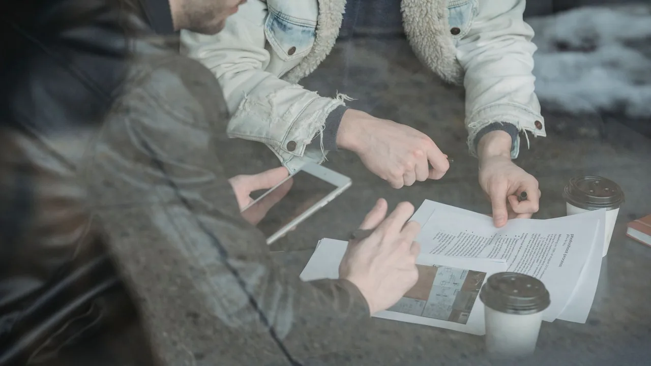 Des mains annotent un document imprimé à côté d'une tablette et de gobelets de café, vue à travers une vitre