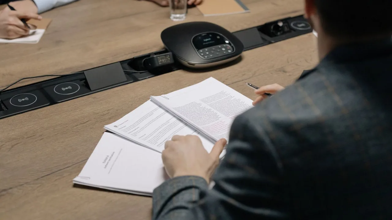 Gros plan sur des mains qui parcourent des documents lors d'une réunion, un téléphone de conférence sur la table