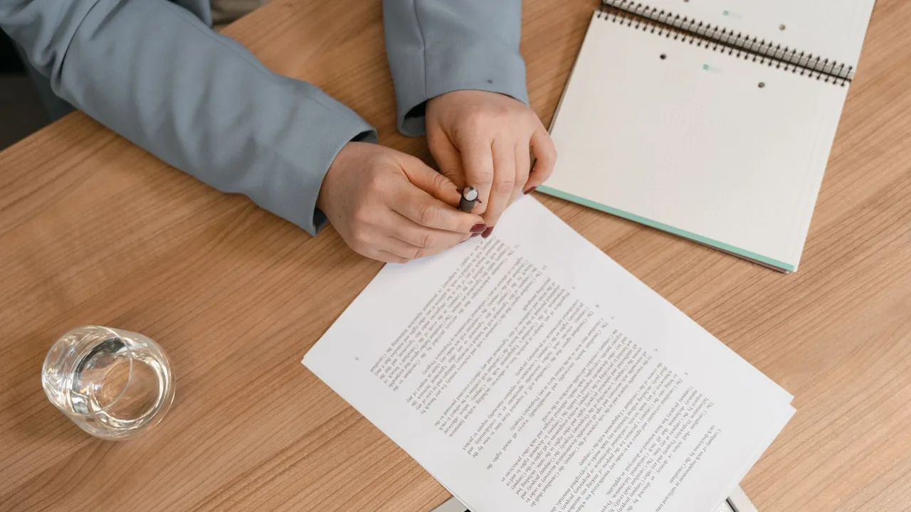 Mains tenant un stylo au-dessus d'un contrat imprimé posé sur un bureau en bois, avec un carnet et un verre d'eau