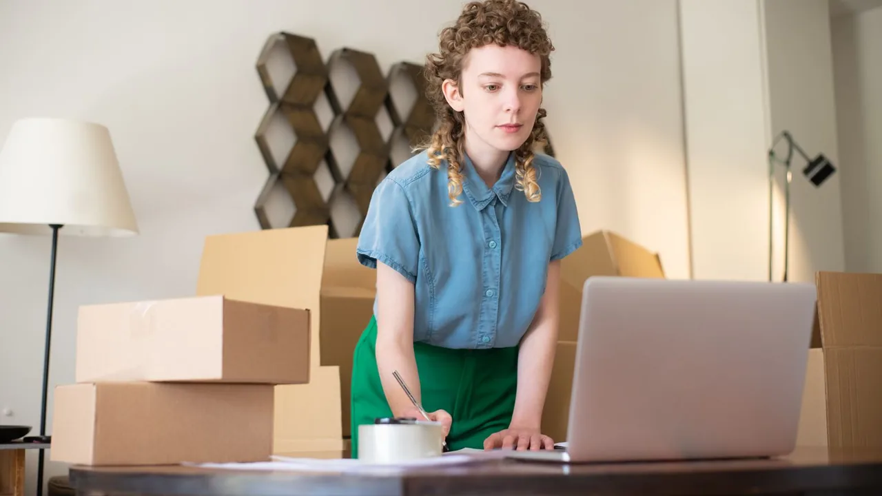 Une jeune femme note quelque chose devant son ordinateur portable, entourée de cartons de déménagement dans un salon