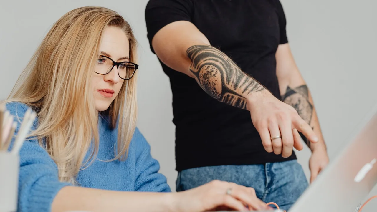 Une femme blonde à lunettes travaille sur son ordinateur portable pendant qu'un collègue tatoué tend le bras vers l'écran
