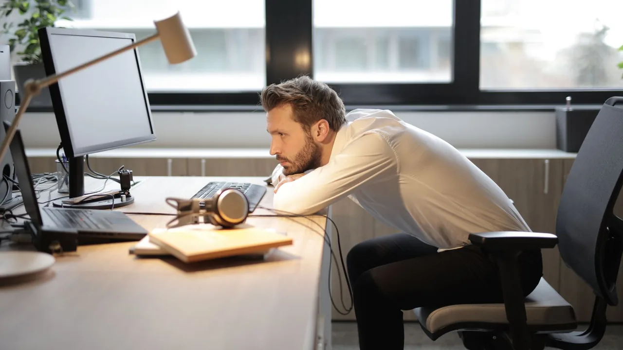 Un homme en chemise blanche, penché sur son bureau, fixe son écran d'un air fatigué