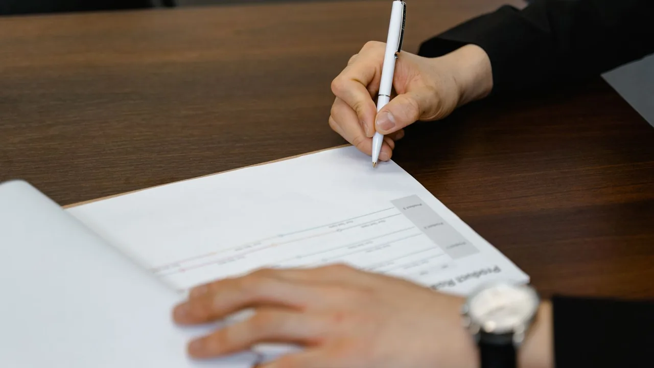 Une main écrit sur un document posé sur un bureau en bois sombre, l'autre main retenant la feuille