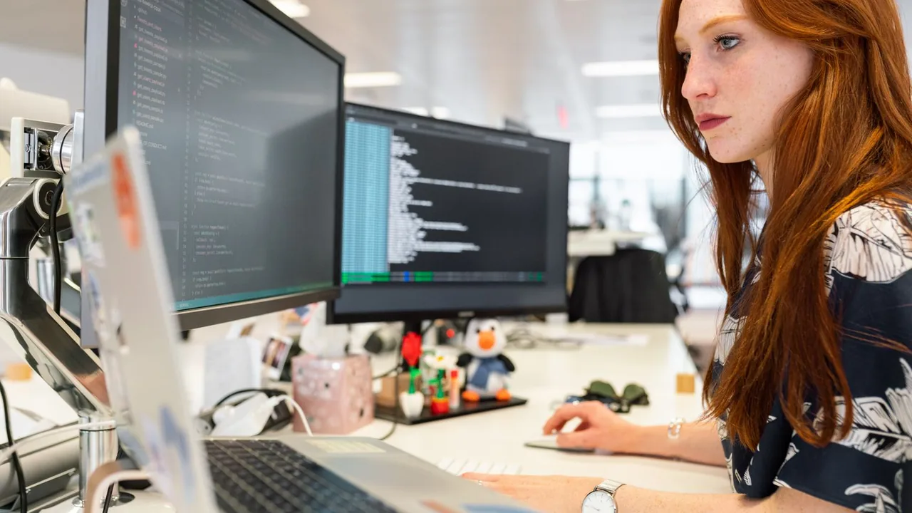 Une développeuse aux cheveux roux, concentrée devant deux écrans de code, dans un bureau décoré de figurines