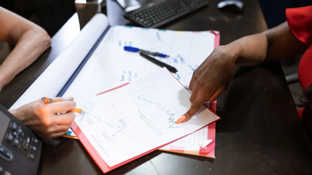 Une main pointe une courbe sur une feuille imprimée posée sur un bureau, stylo et classeur rouge à côté
