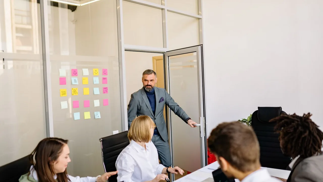 Une équipe réunie dans une salle vitrée, devant une paroi couverte de post-it colorés ; un homme ouvre la porte