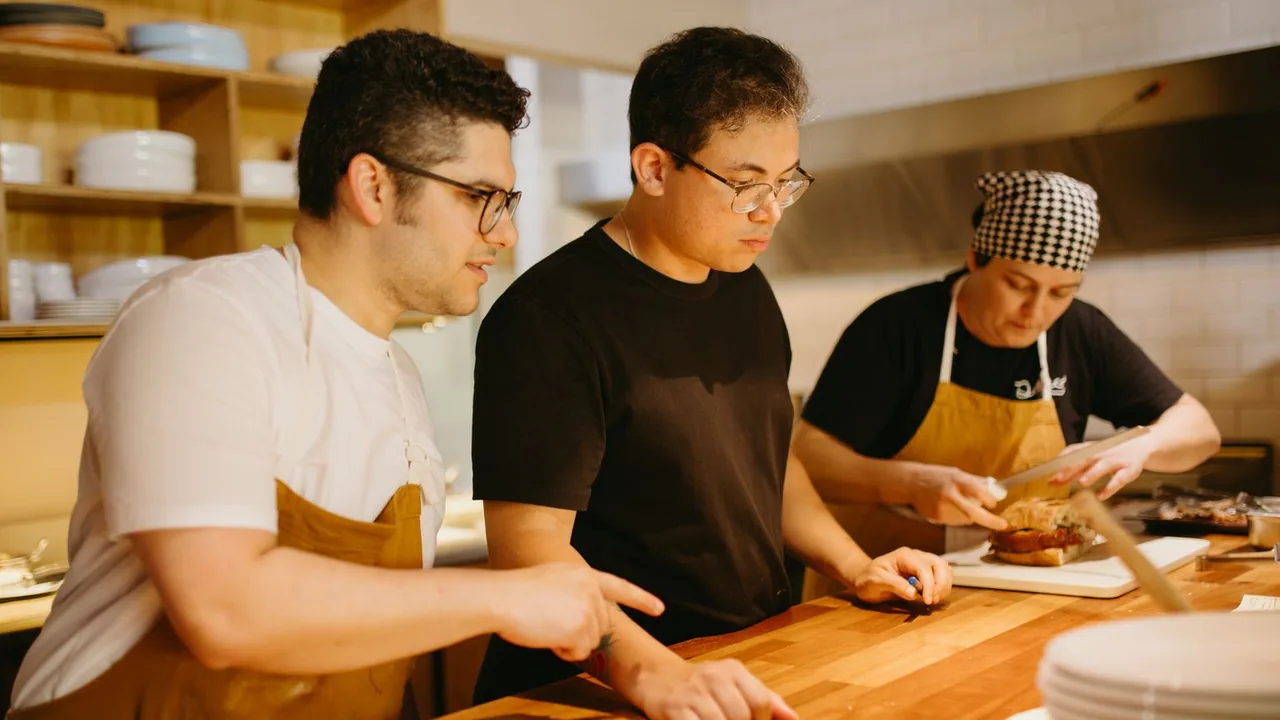 Trois cuisiniers penchés sur un plan de travail en bois, l'un montrant du doigt une préparation