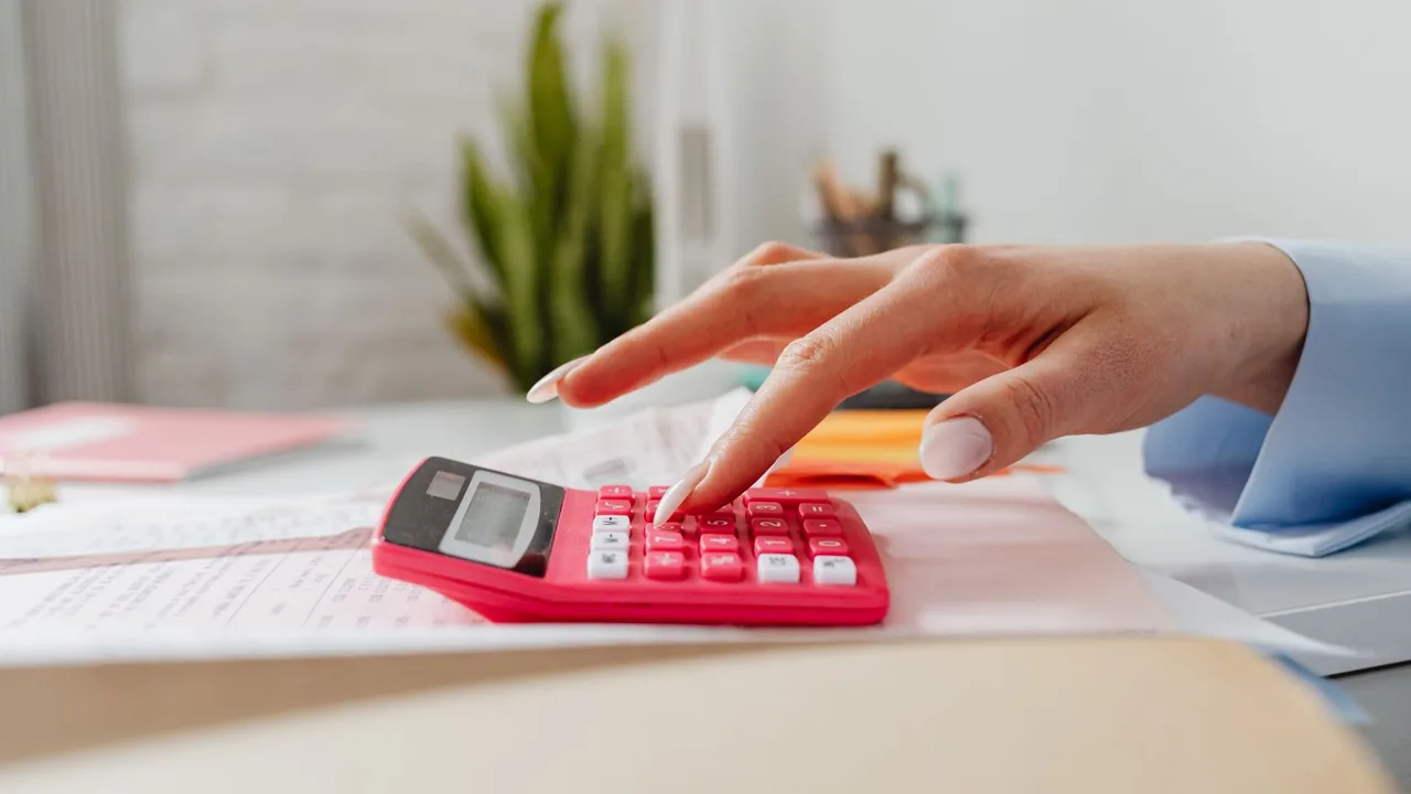 Une main tapote sur une calculatrice rose posée sur une pile de justificatifs, sur un bureau clair