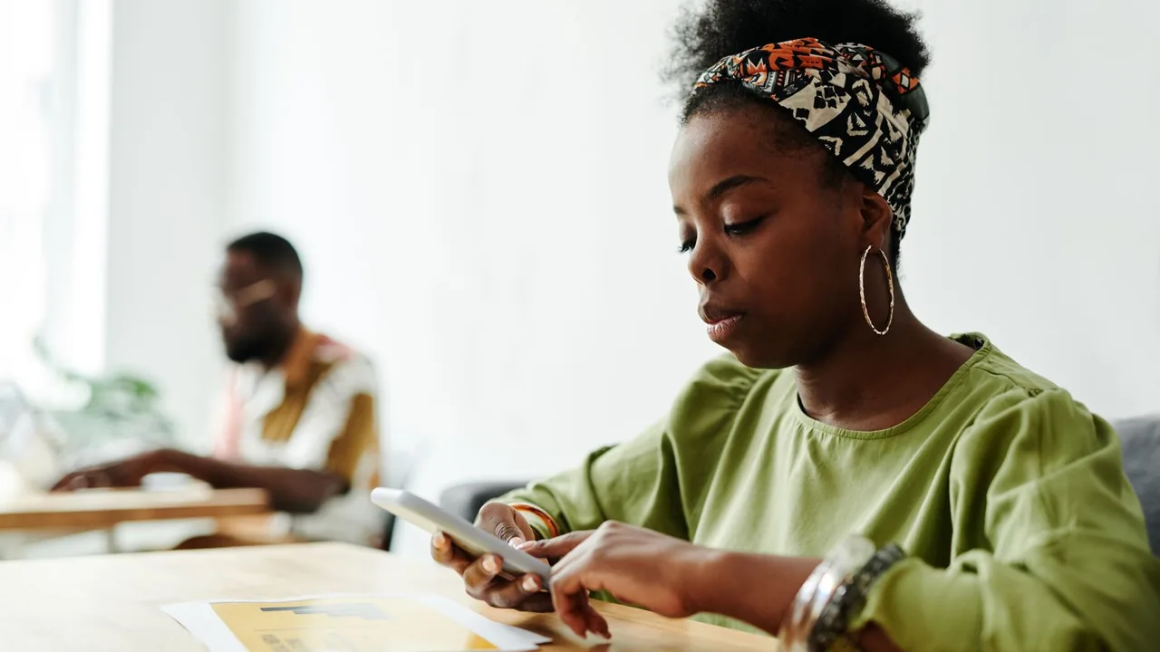 Une femme concentrée utilise son smartphone à une table, un collègue travaille en arrière-plan