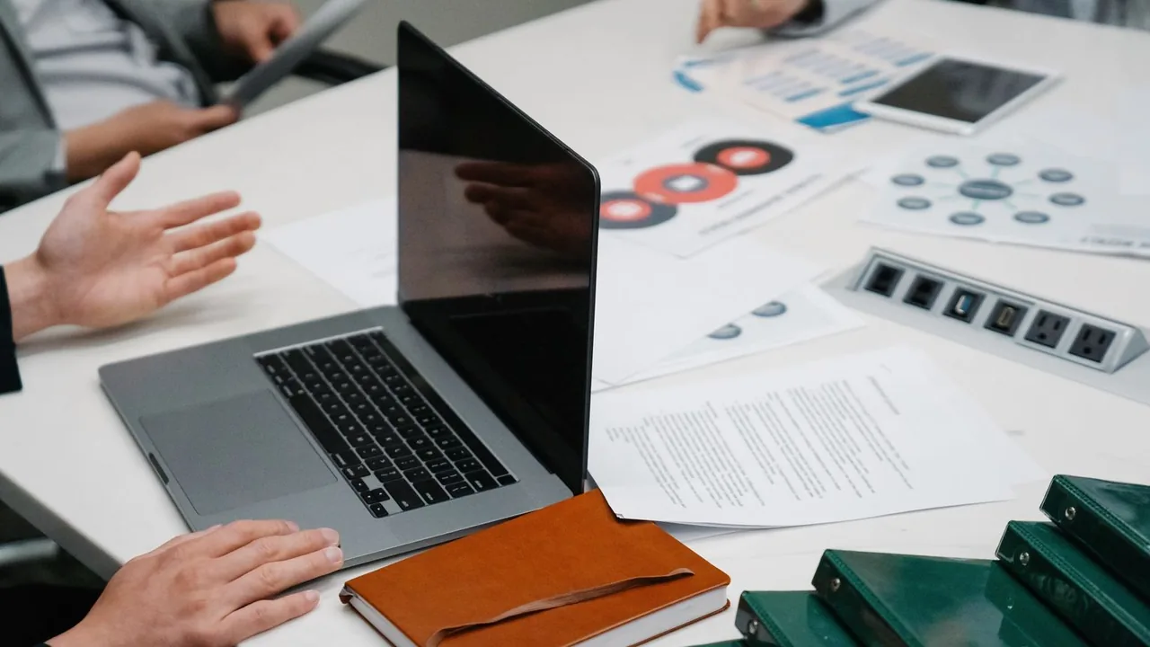 Table de réunion avec un ordinateur portable, des documents et des classeurs, des mains en pleine discussion