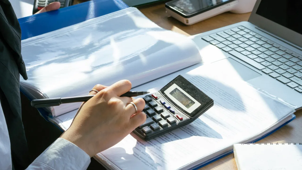 Mains d'une comptable avec calculatrice et stylo sur des dossiers, ordinateur portable et terminal de paiement derrière