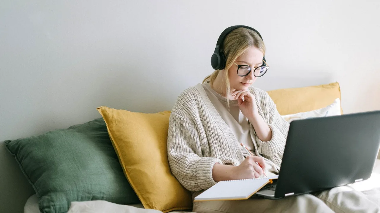 Une apprenante au casque audio suit un cours sur son ordinateur portable, assise sur un canapé, un carnet sur les genoux.