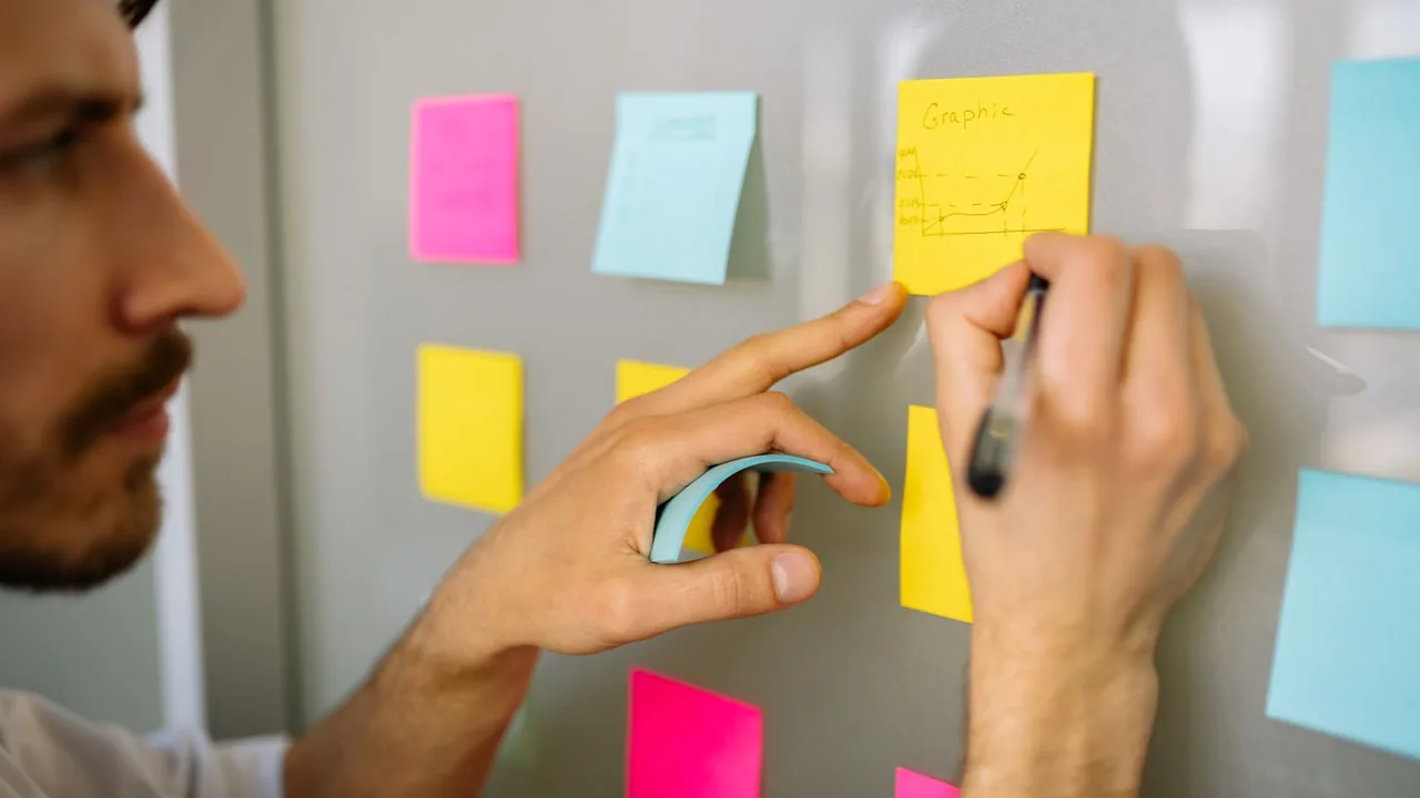 Gros plan sur des mains qui placent et annotent des post-it colorés sur une paroi de bureau.