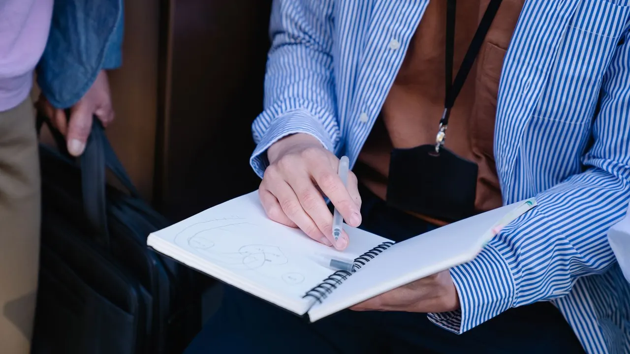 Une personne en chemise rayée, un badge autour du cou, dessine dans un carnet à spirales