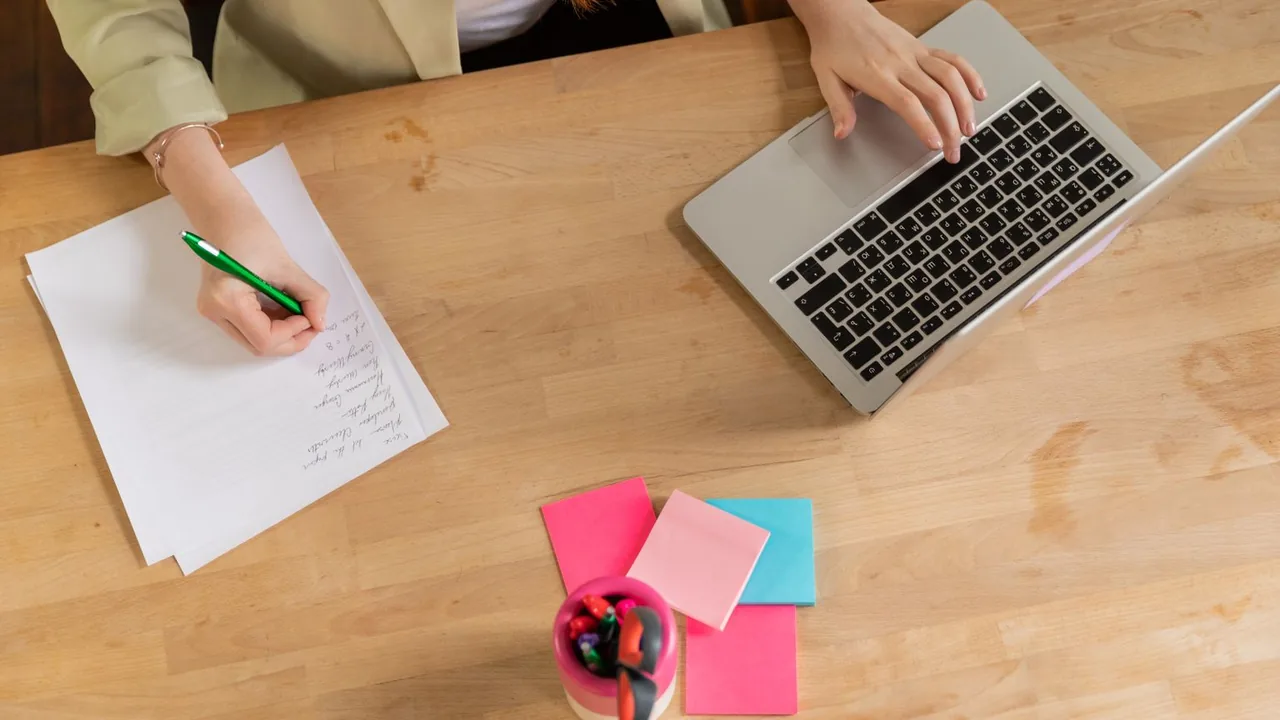 Vue de dessus d'une personne qui écrit sur une feuille pendant qu'elle tape sur un ordinateur portable, sur un bureau en bois
