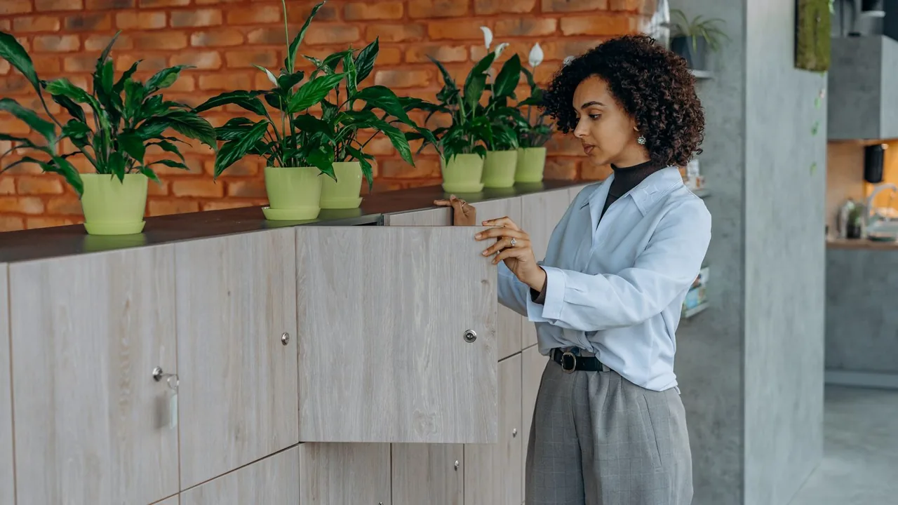 Une femme tient la porte d'un meuble de rangement en bois clair sous une rangée de plantes vertes