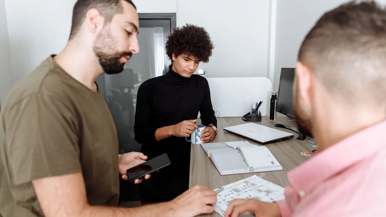 Trois collègues discutent autour d'une table de bureau, carnet et ordinateur portable devant eux