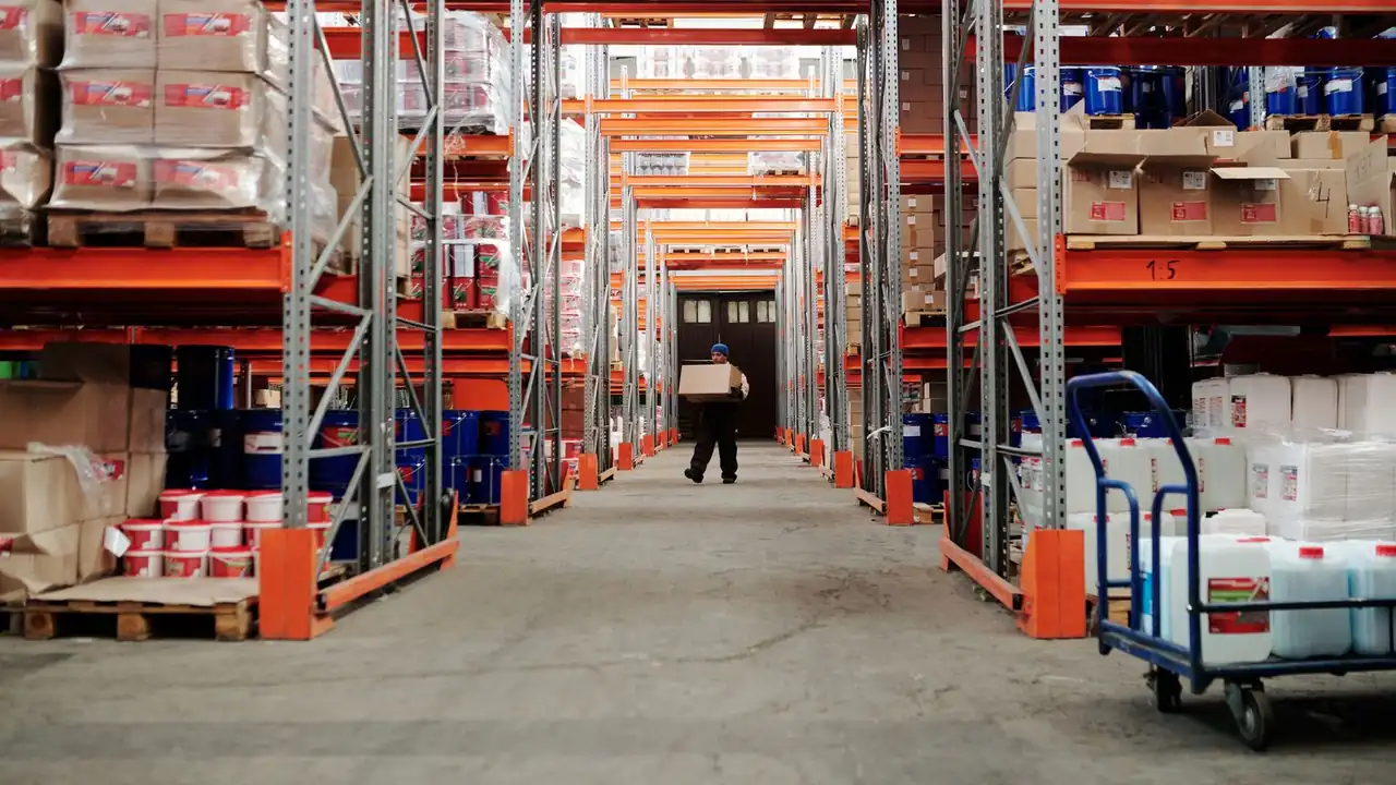 Une longue allée d'entrepôt bordée de rayonnages métalliques chargés de cartons, un employé au loin