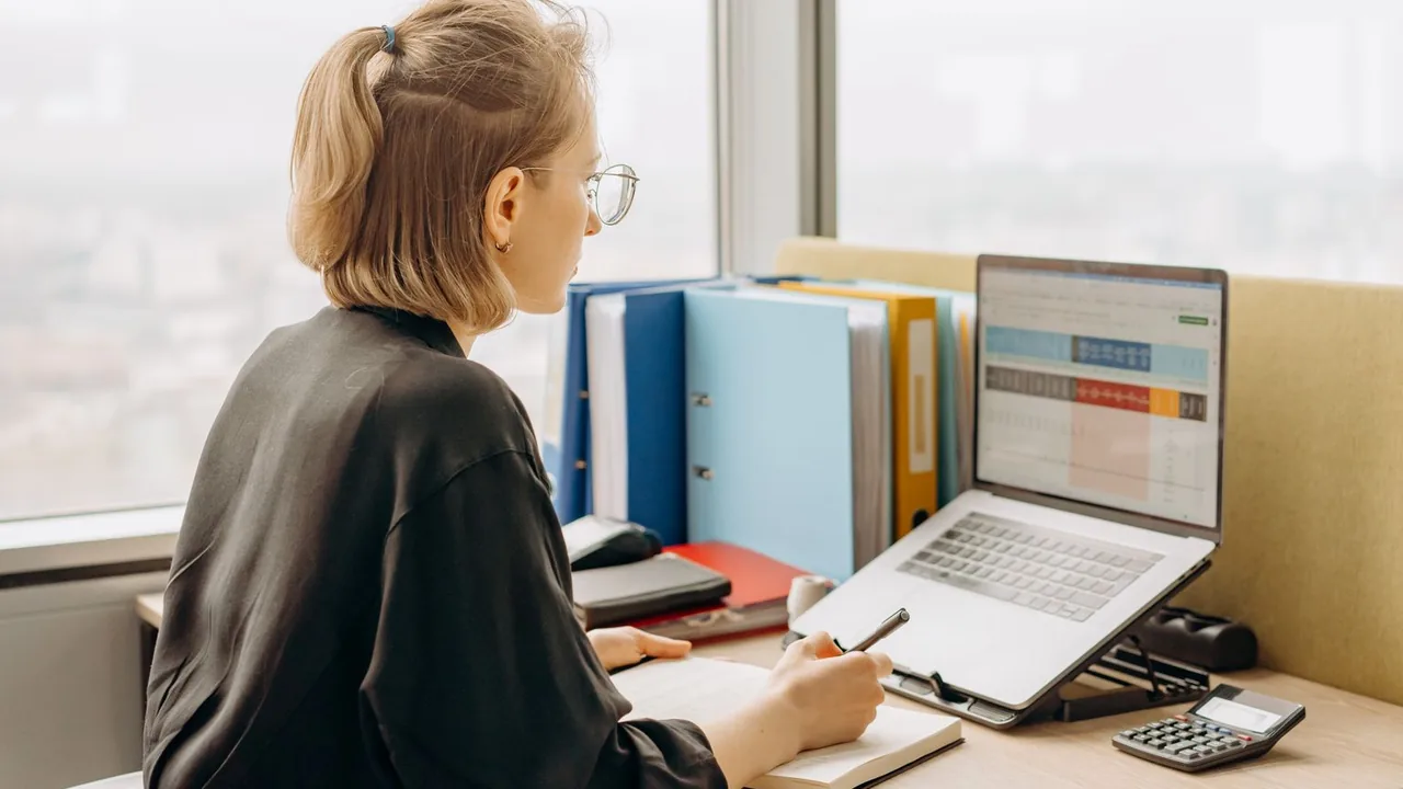 Femme travaillant sur ordinateur portable à son bureau près d'une fenêtre, classeurs à portée de main