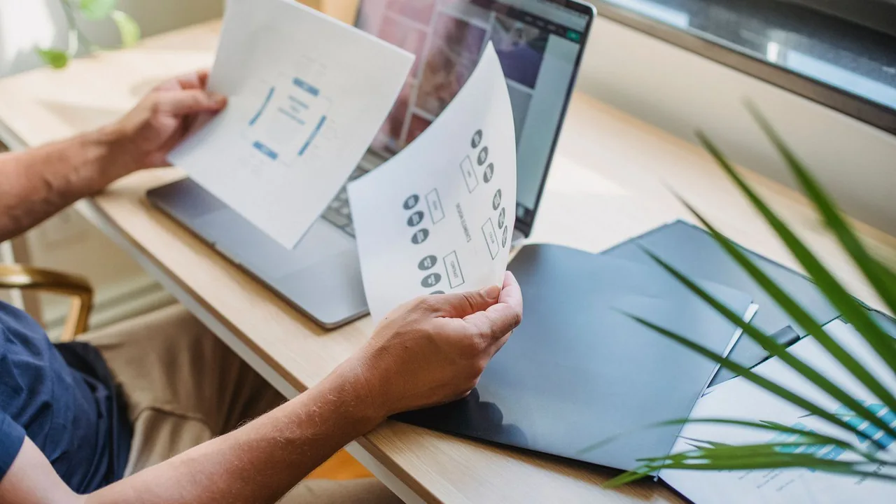 Mains tenant des feuilles imprimées devant un ordinateur portable, plante verte au premier plan