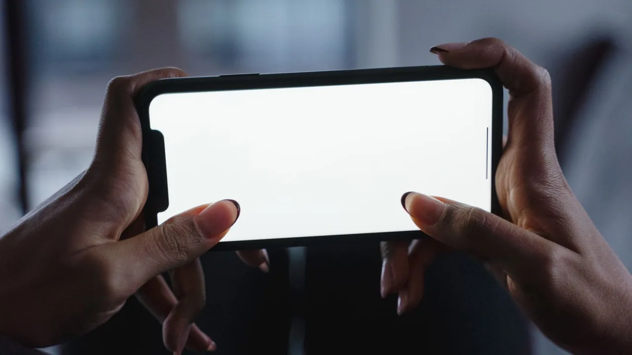 Des mains sombres tiennent un smartphone à l'horizontale, écran blanc, sur fond flou