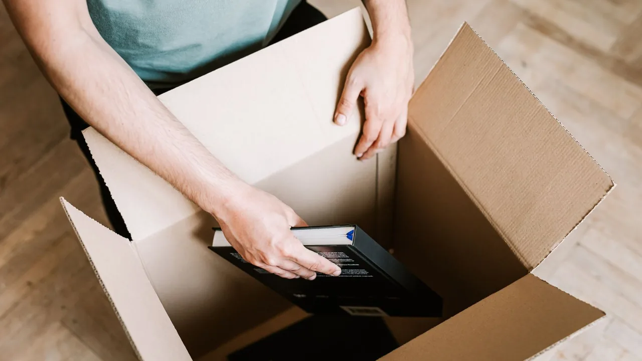 Vue de dessus de mains rangeant un livre dans un grand carton ouvert posé sur un parquet clair