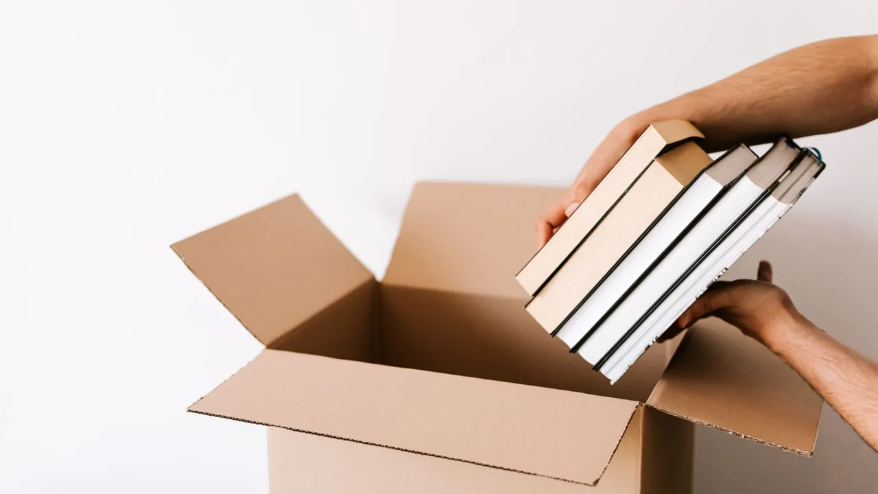 Mains posant une pile de livres dans un carton ouvert devant un mur blanc